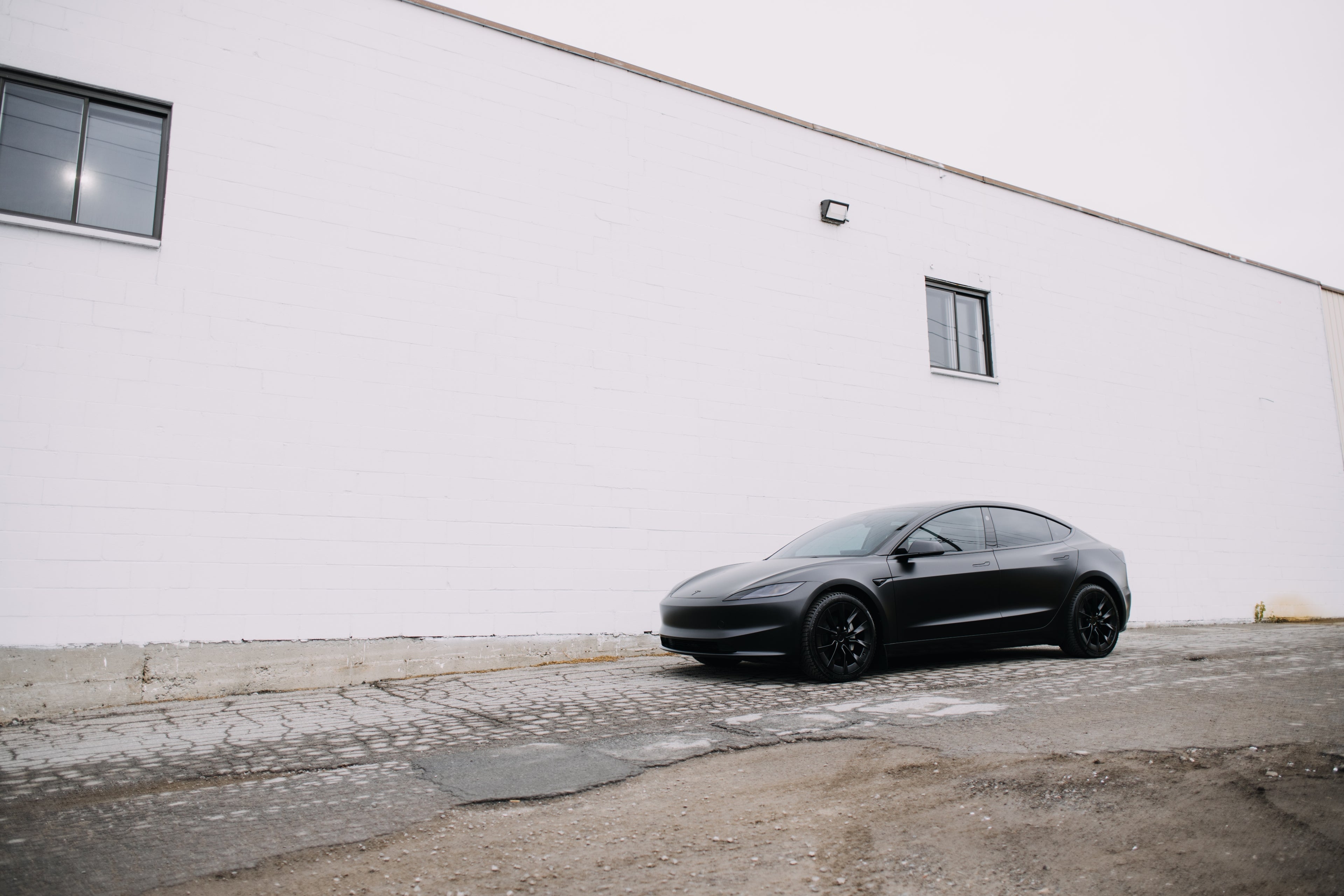 Black Tesla Model 3 parked outdoors in an industrial setting, in front of a white brick wall